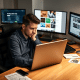 A person sits at a wooden desk, focused on a laptop with two monitors displaying various content in a modern office setting.